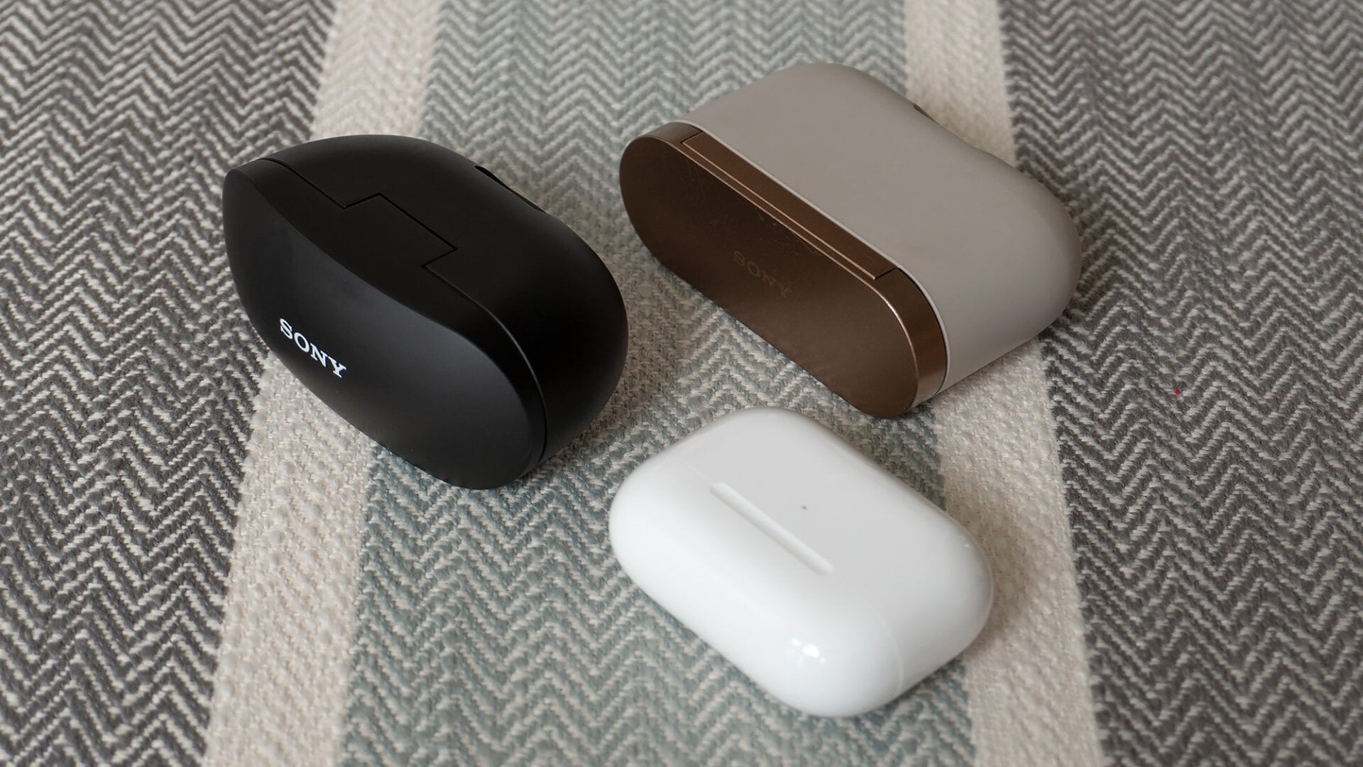 The charging case for the Sony WF-SP800Ns (black) compared to the case for the WF-1000XM3s (tan and gold) and the case for the Apple AirPods Pro (white).