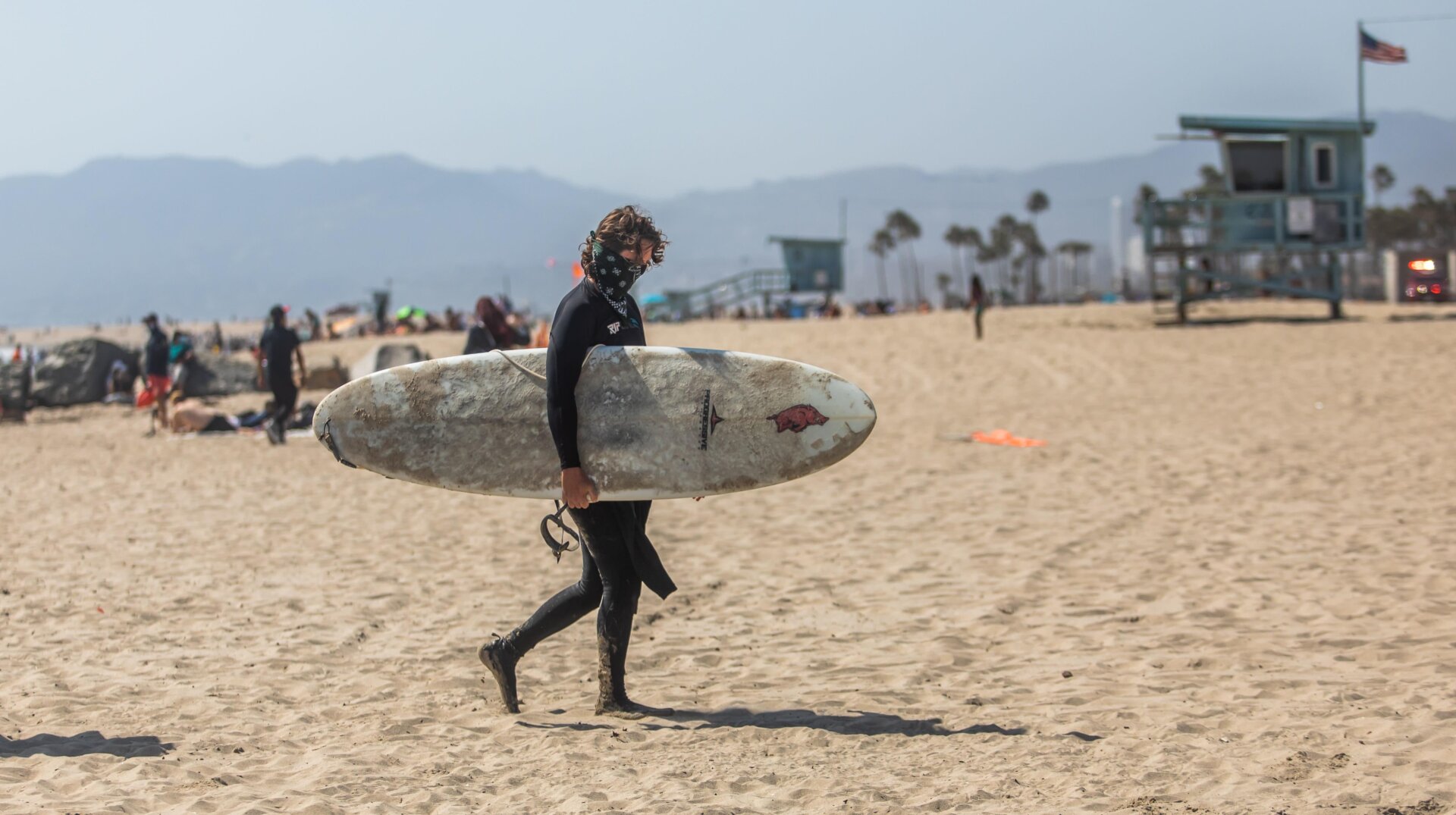 A masked surfer walking at Venice Beach in California during Memorial Day weekend in May.
