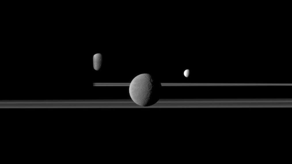 Rhea seen in front of Saturn’s rings. The moons Dione, left, and Enceladus, right, are in the background.