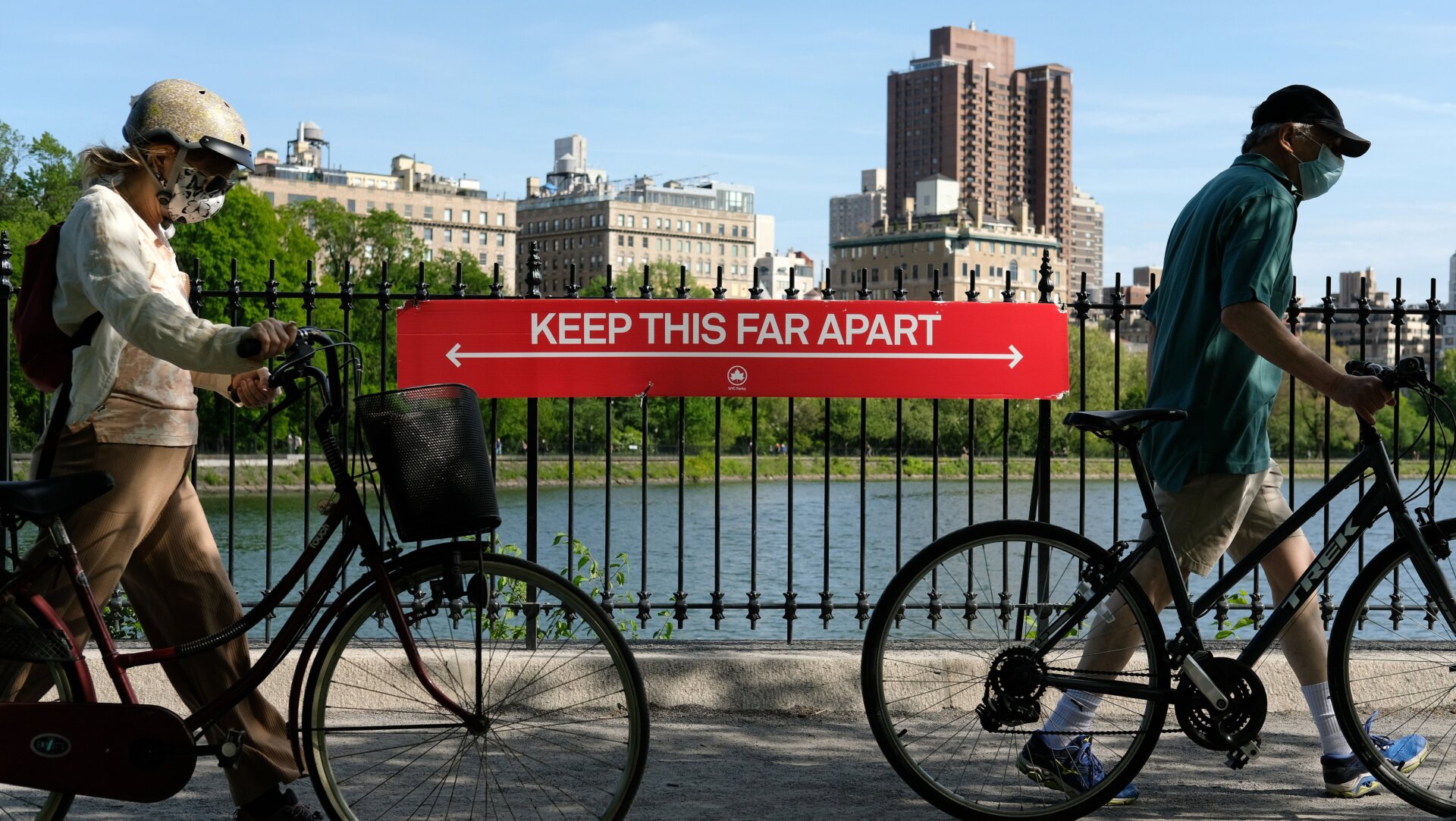 People wearing protective masks walk their bicycles past a social distancing sign reading “KEEP THIS FAR APART” at Jacqueline Kennedy Onassis Reservoir in Central Park during the coronavirus pandemic on May 17, 2020 in New York City.