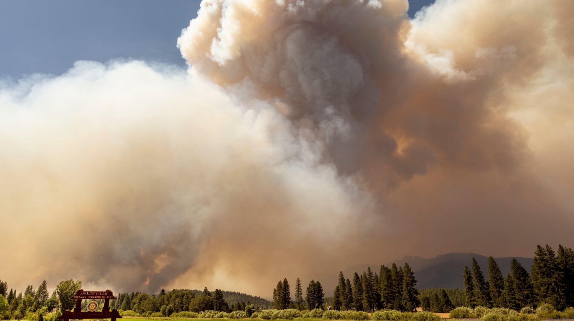 A Greenville, California welcome sign stands in front of a billowing smoke plume as the Dixie fire burns on August 3, 2021.