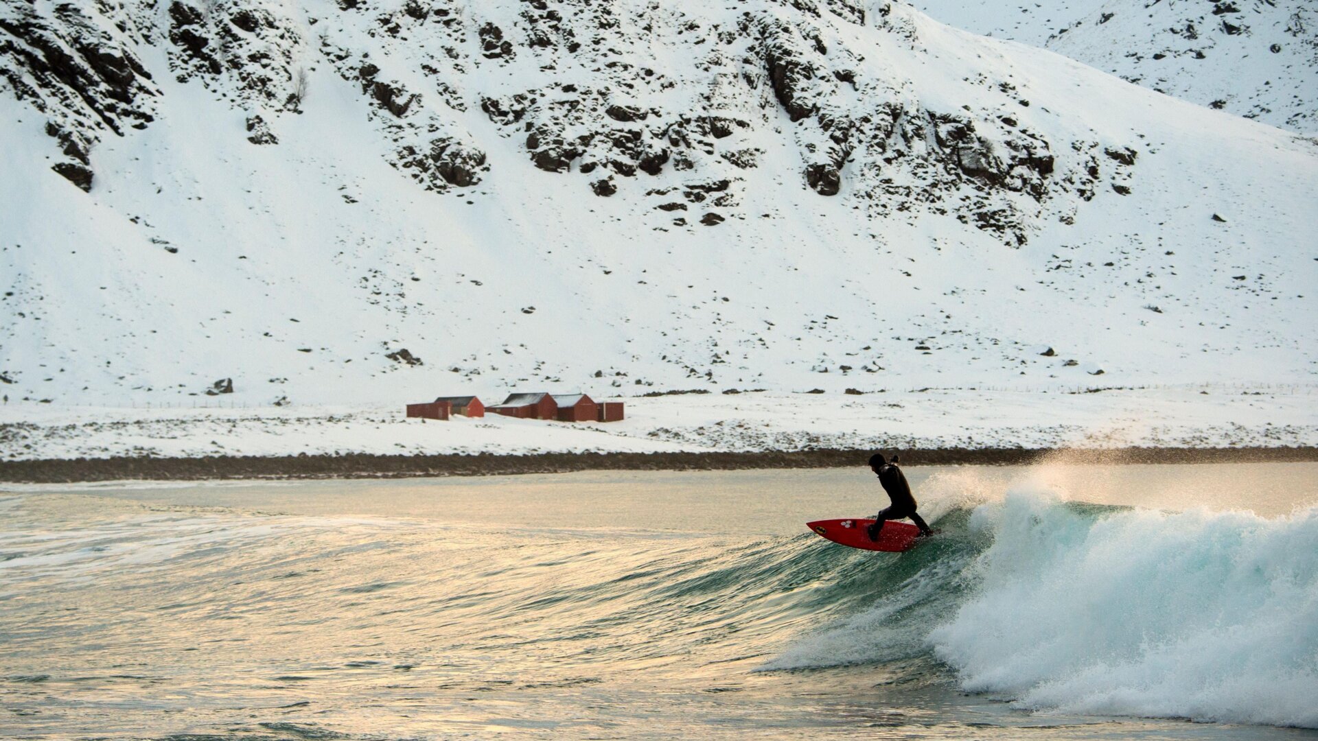 A surfer rides a wave at the snowy beach of Unstad, in Lofoten Island, Arctic Circle.