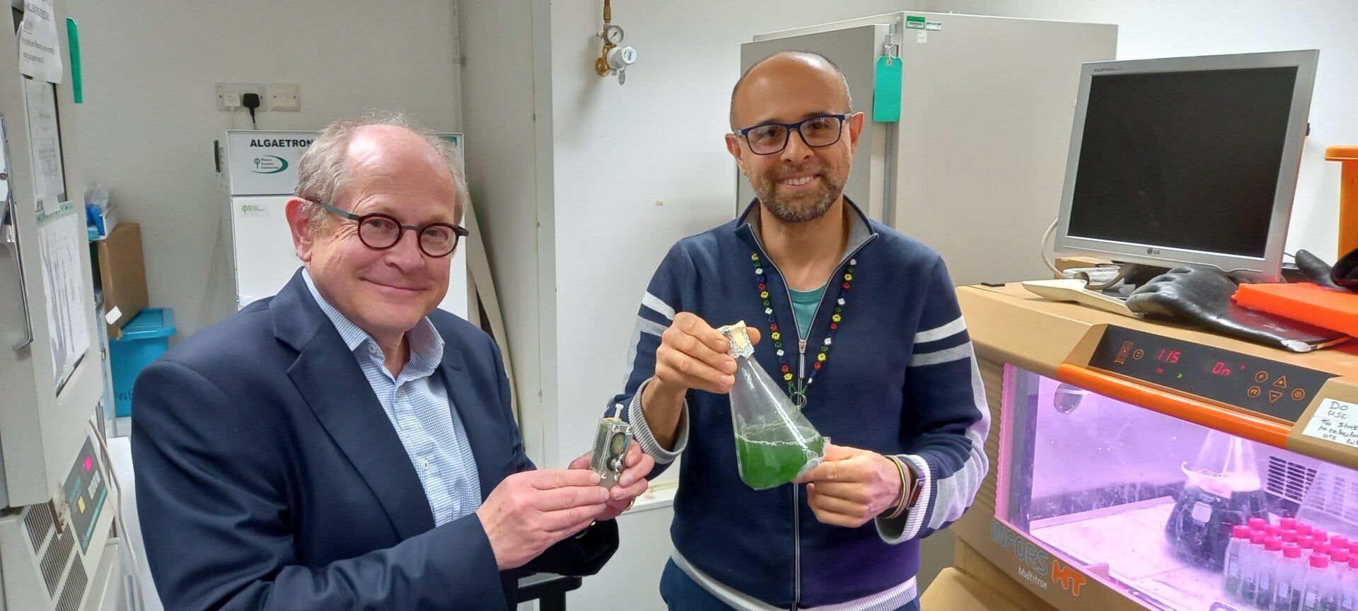 Scientists Chris Howe (left) and Paolo Bombelli (right) holding their algal power unit and a flask of cyanobacteria solution.