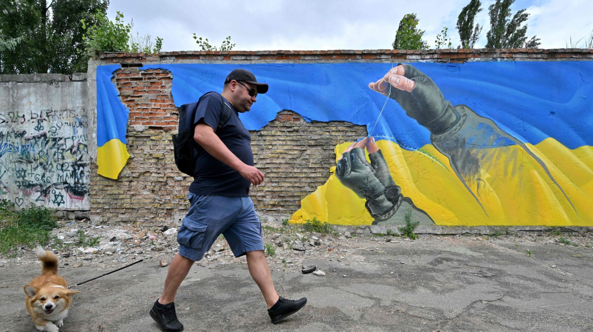 A man walks with his dog past a mural by street artist Sasha Korban depicting hands of a military man sewing together parts of the Ukrainian flag, in Kyiv on June 14, 2022.