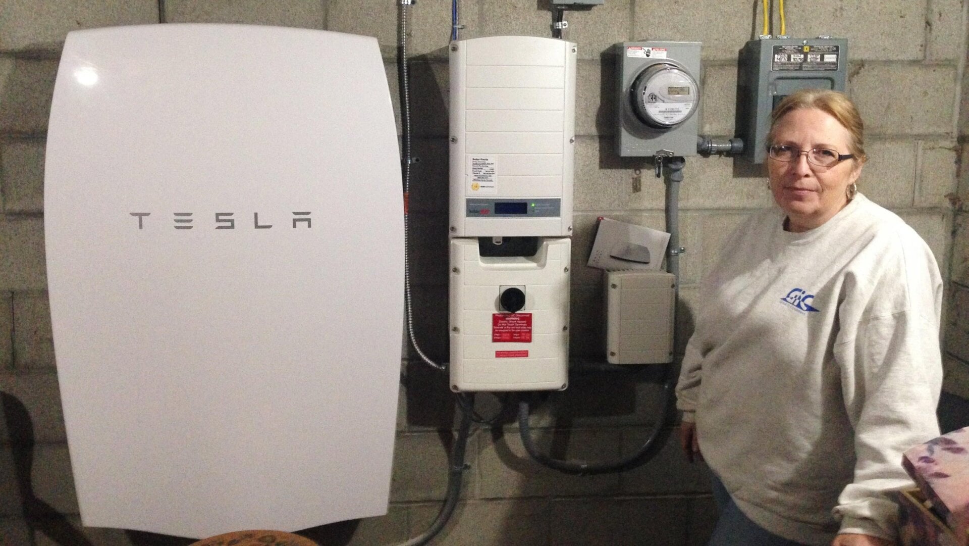 A customer in Vermont stands next to a Tesla Powerwall battery and inverter which she uses to store solar energy.
