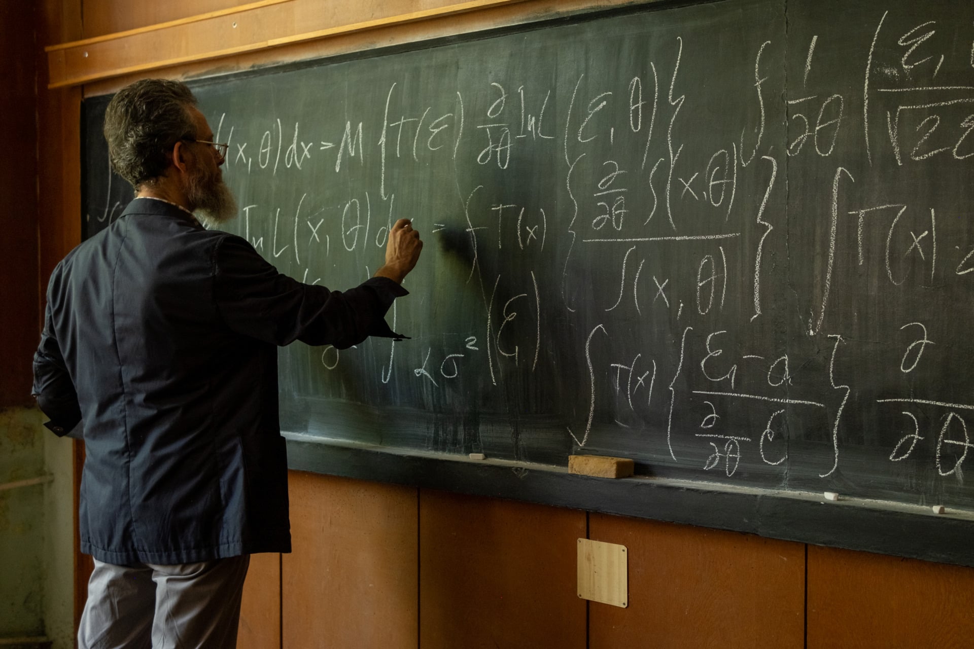 A Teacher With A Long White Beard, Big Gray Hair, And Glasses Is Wearing A Checkered Shirt And Sports Jacket In Front Of A Blackboard With Complex Formulas Written On It.