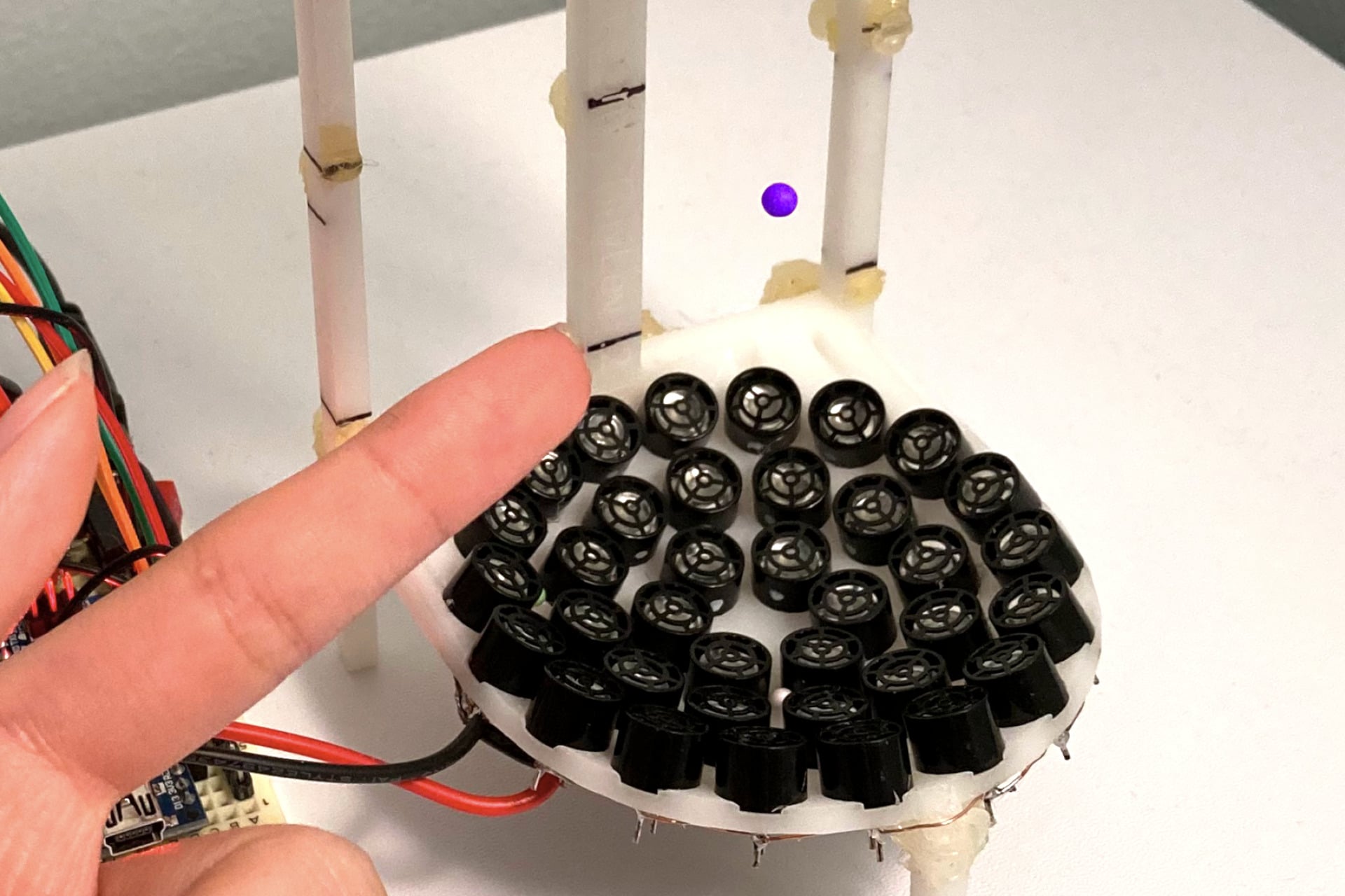 A bead (purple) is suspended in mid-air by sound waves emanating from (black) circular speakers arranged in a six-inch-tall 3D-printed frame.