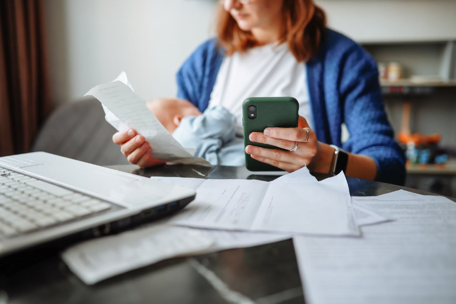 Person filing taxes, holding phone in one hand and receipt in the other, laptop and papers in front of her