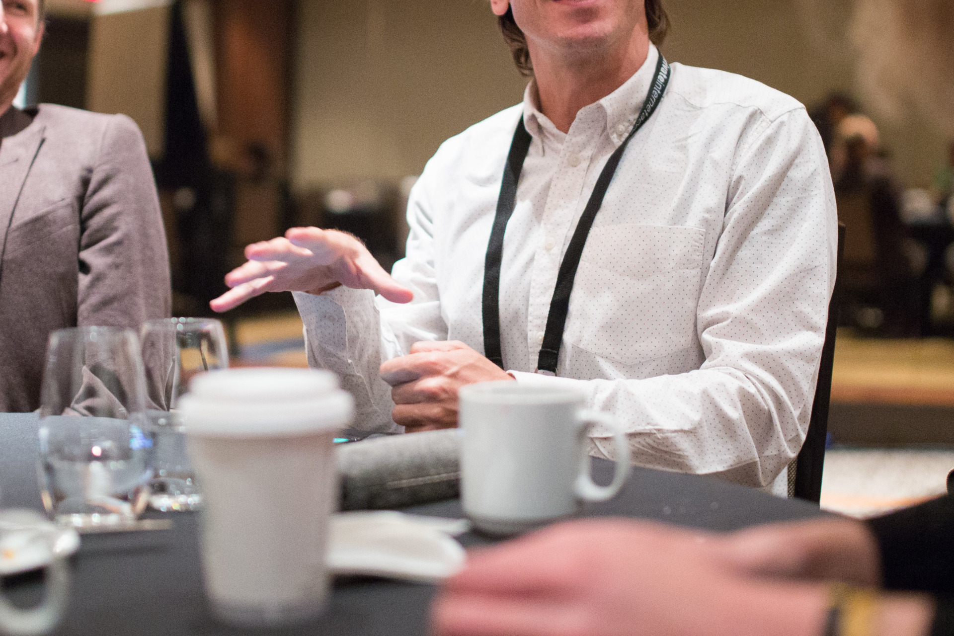 Above, men with lanyards around their necks talk jovially surrounded by coffee and water in a conference room.