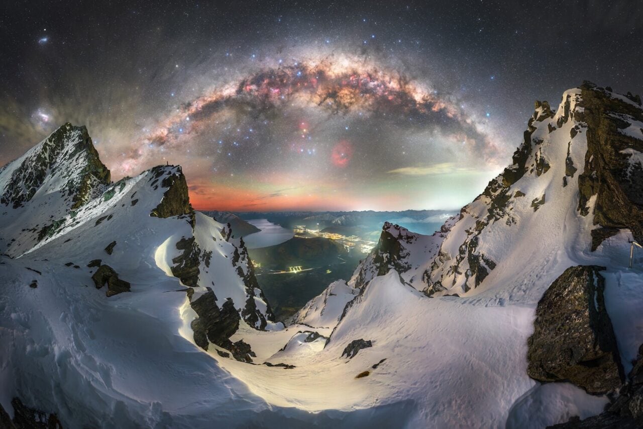 The winter Milky Way arch sets above the rugged Remarkables Range in the Southern Alps, while the lights of Queenstown, New Zealand, glow far below.