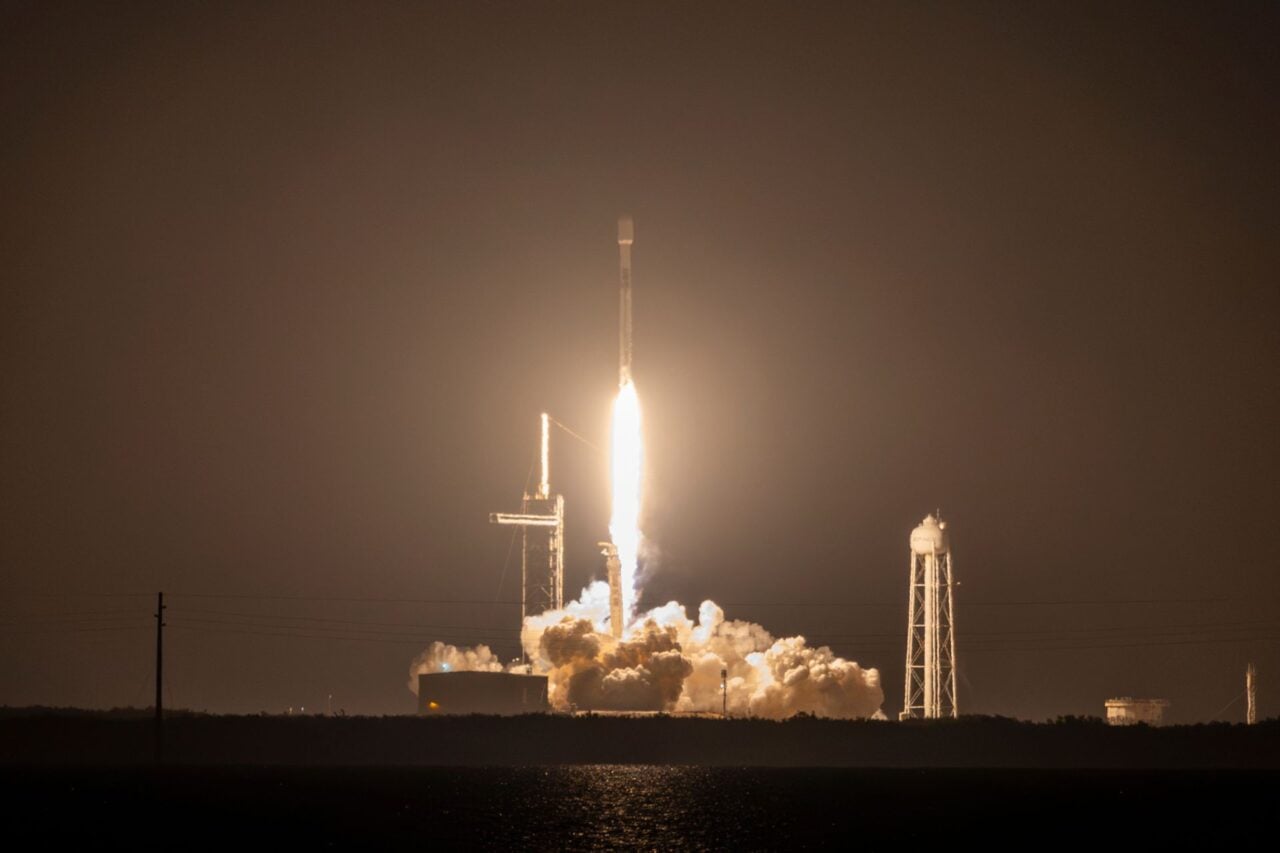 In the distance, a Falcon 9 rocket blasts off, illuminating the night sky