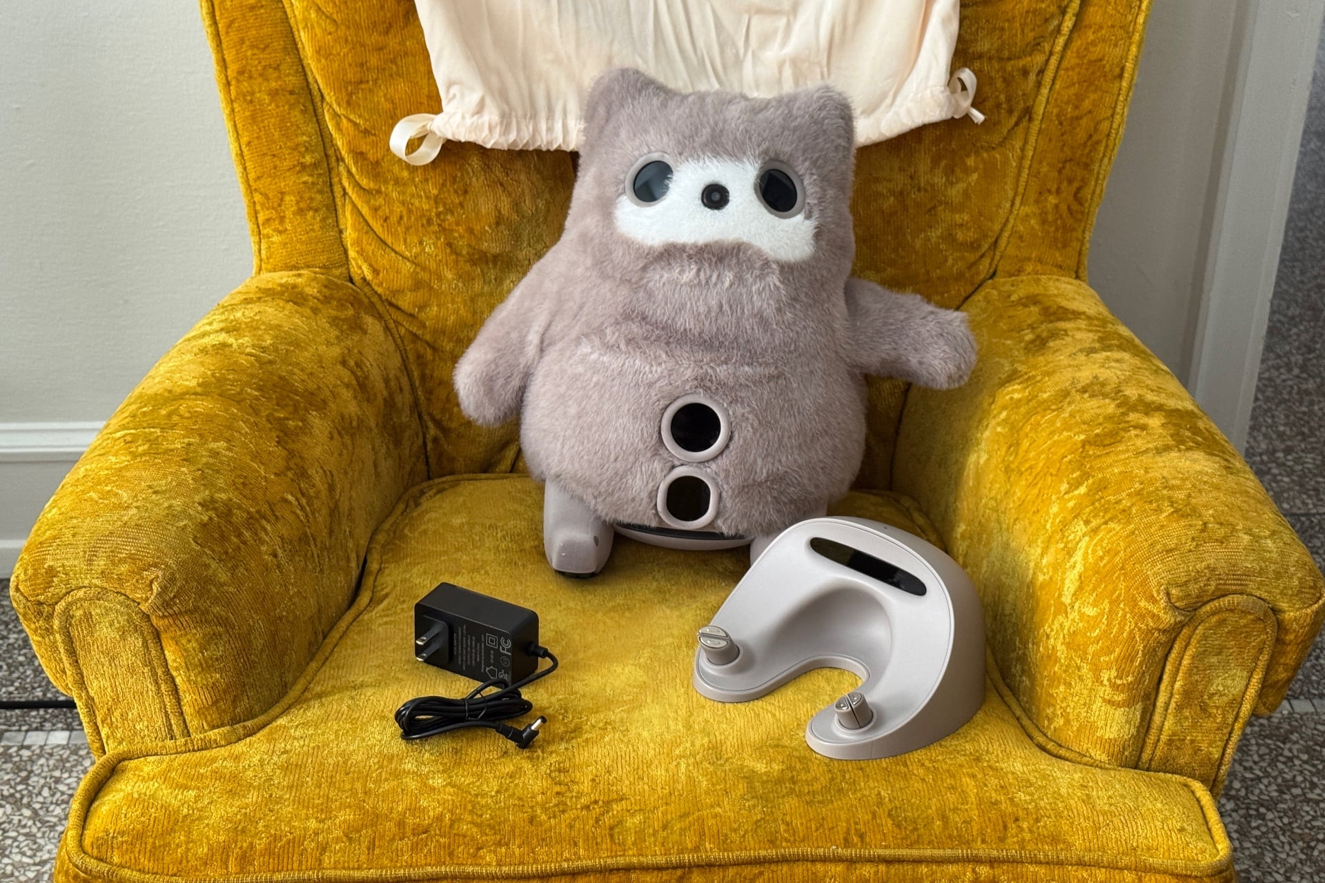 The Switchbot Kata Friends pictured sitting on a chair with its eye displays off, next to its dock and power adapter.