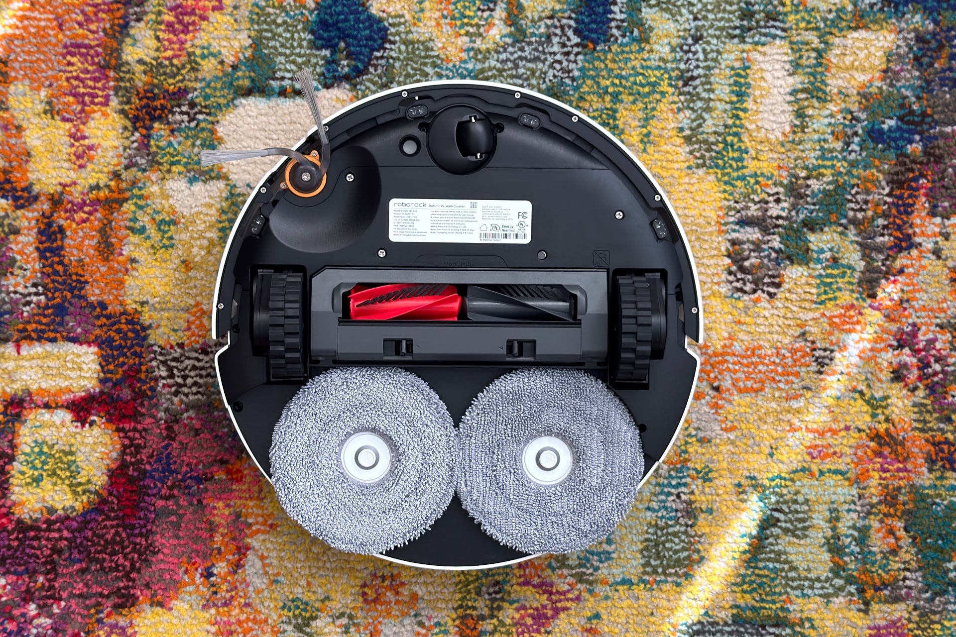 Photo of the bottom of the Roborock Qrevo 2 Pro, with its mop pads installed. Its two-piece roller brush is red and gray, mop pads are light gray, and its dual-brush side brush has an orange circle around it. Otherwise, the bottom is black plastic.