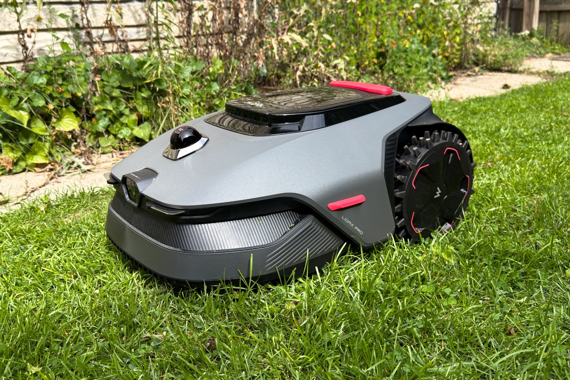 Mova Lidax Pro 800 three-quarter shot as it sits in grass. It's a grey and black mower that's wedge-shaped, with two large, knobby back wheels. Front wheels aren't visible. There is a camera port in the front, a skirt-like bumper below that, and a LiDAR sensor just behind the camera on top.