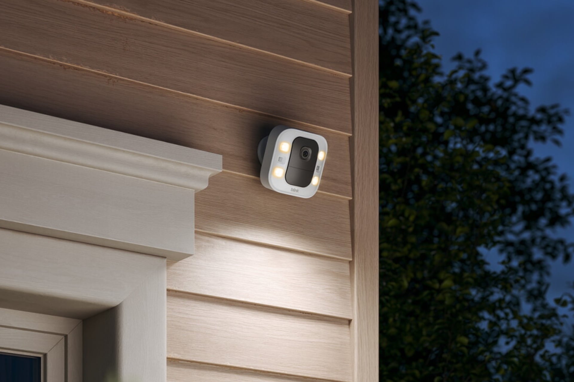 Blink Spotlight Cam 2k Plus mounted on a wall. It's square and white with a black verticle rectangular section in the middle, flanked by sets of two LED lights on either side. All of the corners of the shapes are rounded.
