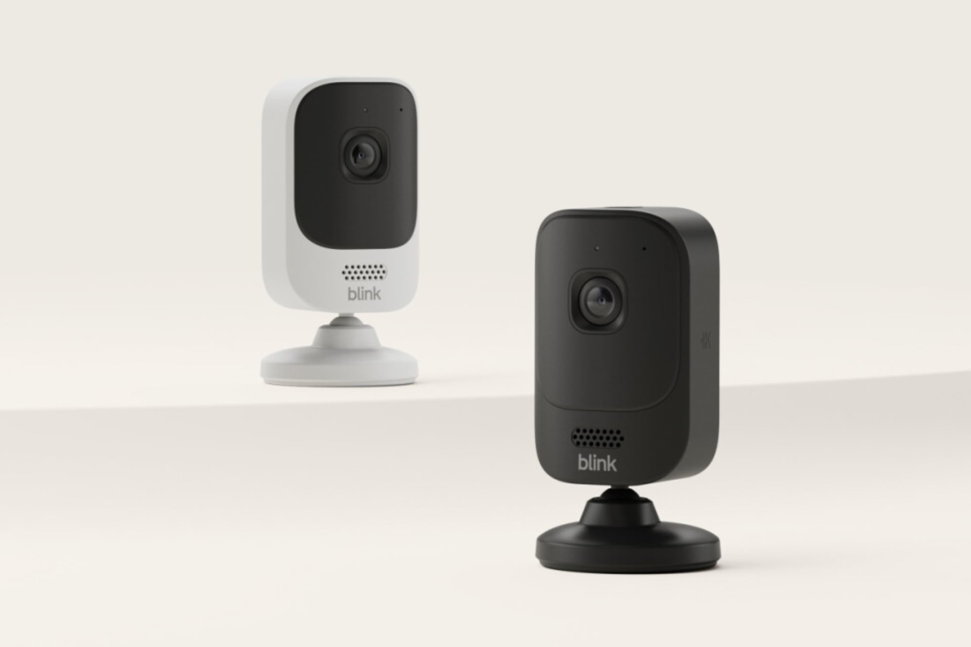Two Blink Mini 4K_ cameras, one in black and the other in white. The two are taller than wide, rectangular with rounded corners, and connected to a squat base with a pivoting ball joint.